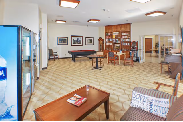 A spacious common area with patterned carpet flooring, a vending machine on the left, a pool table against the far wall, a wooden bookshelf filled with books, several chairs and tables, framed pictures on the wall, and a cushioned armchair with a pillow featuring words like 'LOVE' and 'GIVING'.