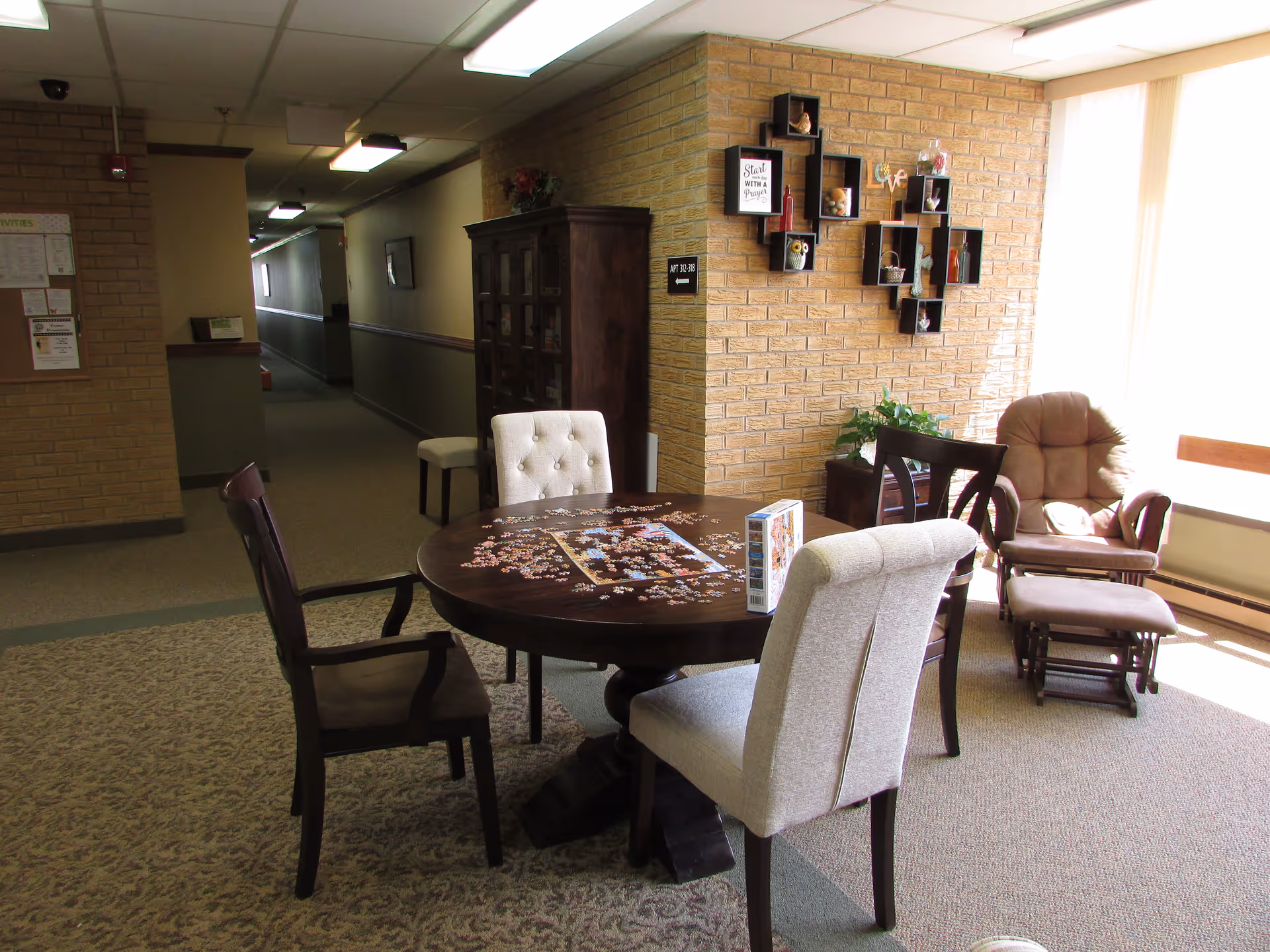 Sunlit communal seating area with a round table mid-assembly jigsaw puzzle, several chairs and a recliner by a brick accent wall.