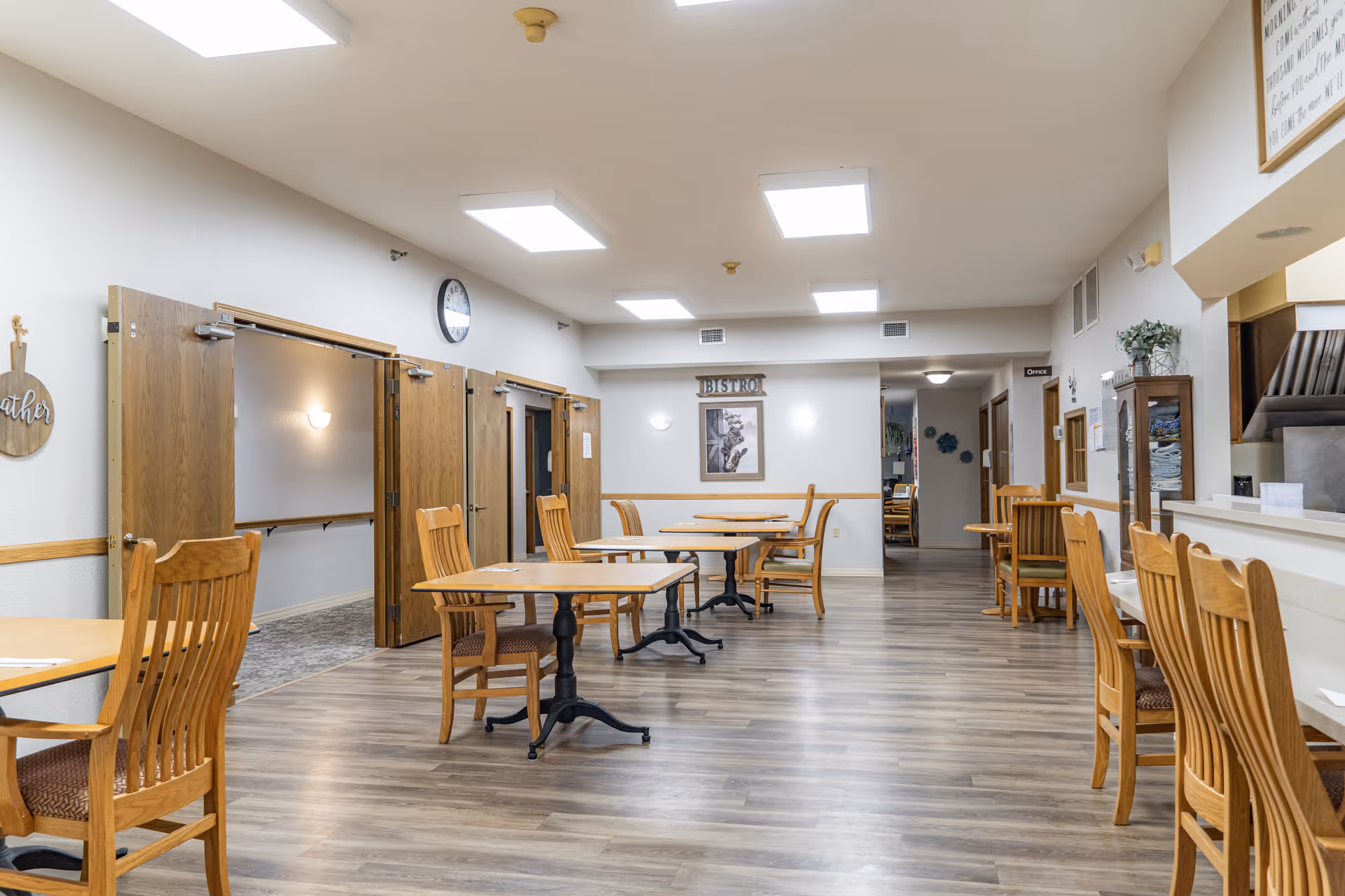A dining area in a senior living facility with wooden tables and chairs arranged neatly on a wood-patterned floor. The room has bright ceiling lights, a clock on the wall, and a sign that reads 'BISTRO'. There is a counter with high chairs on the right side and open double doors on the left leading to another room.