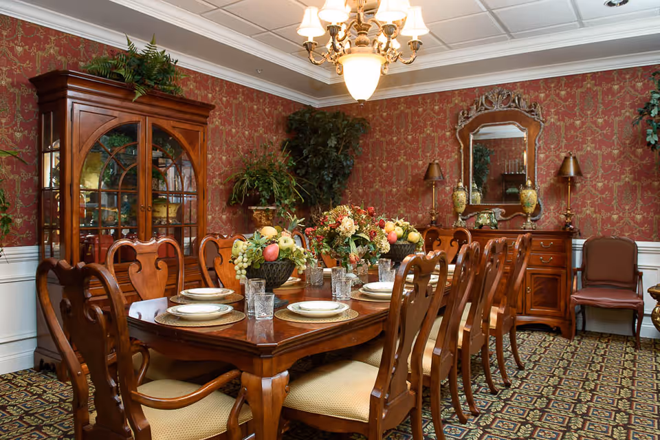 A formal dining room with a large wooden dining table set with plates, glasses, and decorative fruit bowls. The room features ornate red wallpaper, a chandelier, a wooden china cabinet, a sideboard with a mirror, and upholstered chairs.