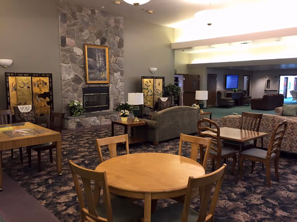 A cozy common area in an assisted living facility featuring multiple wooden tables and chairs arranged on a patterned carpet. In the background, there is a stone fireplace with a framed painting above it, flanked by decorative folding screens and potted plants. Several sofas and armchairs are arranged for seating, with table lamps on side tables providing warm lighting.