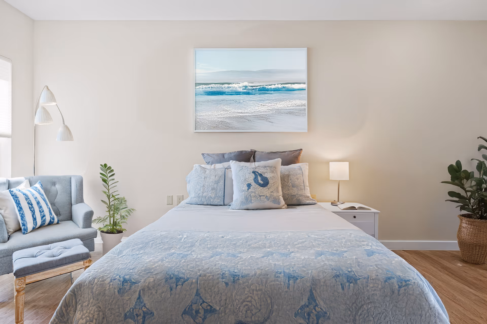 A neatly made bed with light blue and white bedding and multiple pillows in a bedroom. Above the bed hangs a framed picture of ocean waves. To the left of the bed is a light blue armchair with a matching ottoman and a floor lamp with three white lampshades. There are two potted plants, one next to the armchair and one in a woven basket on the right side of the bed. A white nightstand with a small lamp and an open book is on the right side of the bed. The room has light-colored walls and wooden flooring.