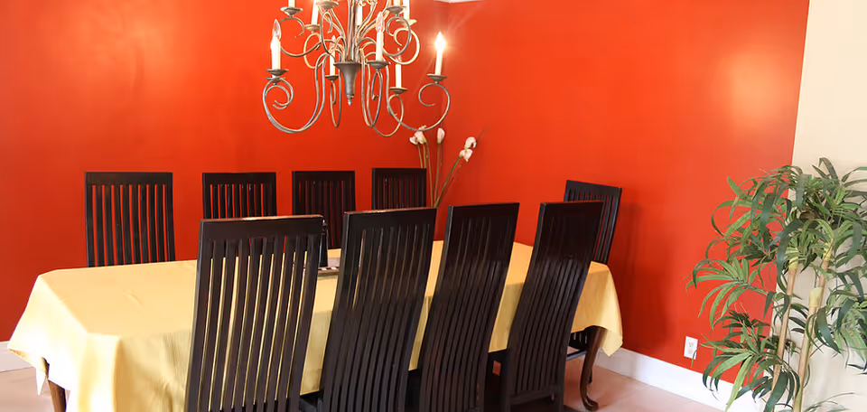 Dining room with a long table covered by a yellow tablecloth, dark wooden chairs, a chandelier, a red accent wall, and a potted plant.