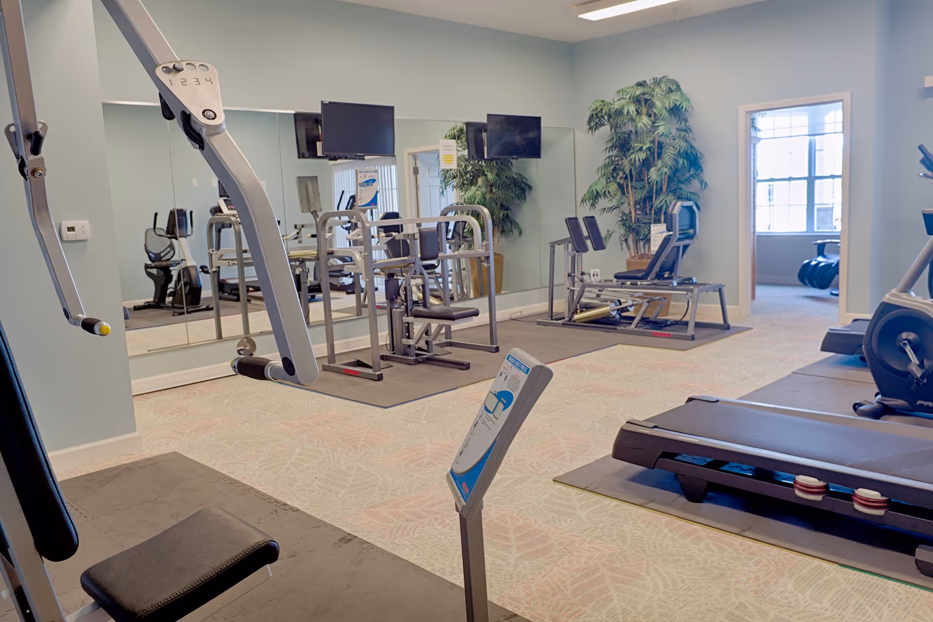 A fitness room with various exercise equipment including a treadmill, weight machines, and stationary bikes. The room has light-colored walls, a large mirror on one side, and a potted plant near the back. There is a windowed room visible through an open doorway.