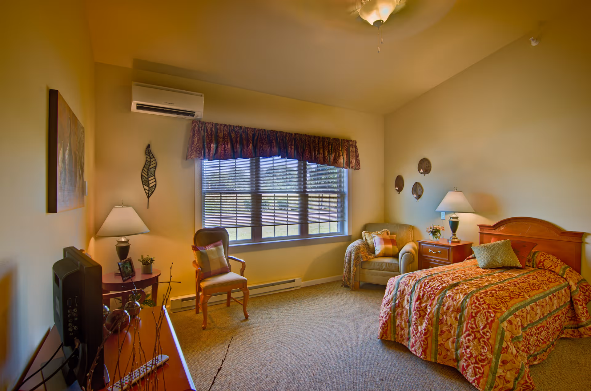 A cozy bedroom in an assisted living facility featuring a single bed with a patterned bedspread, a wooden nightstand with a lamp and flowers, an armchair with a throw blanket and pillow, a wooden chair with a cushion, a window with blinds and a valance, a ceiling fan, and a TV on a wooden dresser.
