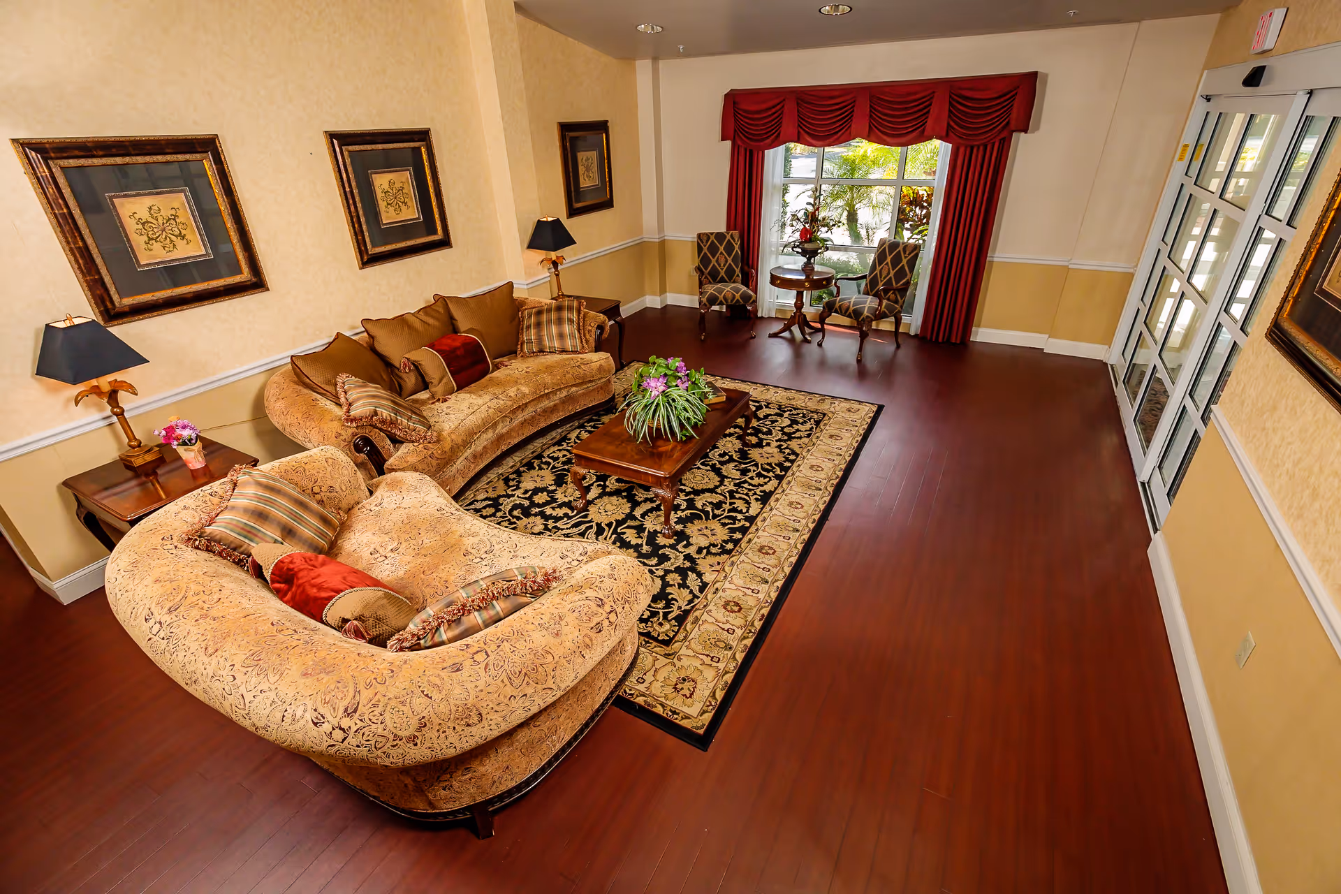 A cozy living room area with two curved beige patterned sofas adorned with various cushions, a wooden coffee table with a floral centerpiece on a decorative rug, two side tables with lamps, framed artwork on the walls, and two upholstered chairs with a small round table near a window with red drapes.