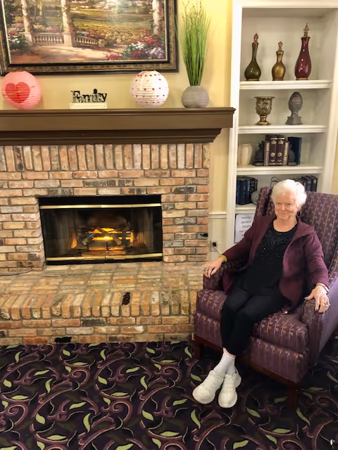 An elderly woman with white hair sitting in a purple patterned armchair next to a brick fireplace with a fire burning. The fireplace mantel is decorated with two paper lanterns, a small sign that says 'Family', and a potted plant. Behind her is a white built-in bookshelf with decorative items and books. The room has a patterned carpet and a framed painting above the fireplace.