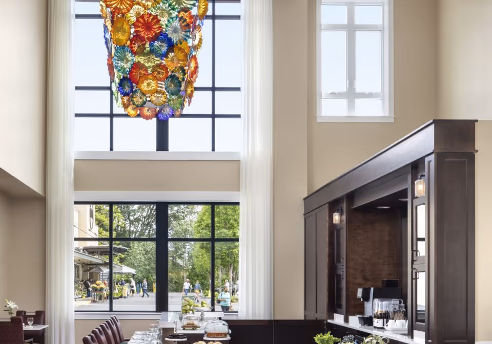 Interior view of a senior living facility dining area with tall windows letting in natural light. A colorful glass chandelier hangs from the ceiling. There is a dark wooden counter with coffee machines and glassware on the right side, and tables with chairs on the left side. Outside the windows, people can be seen walking near greenery.