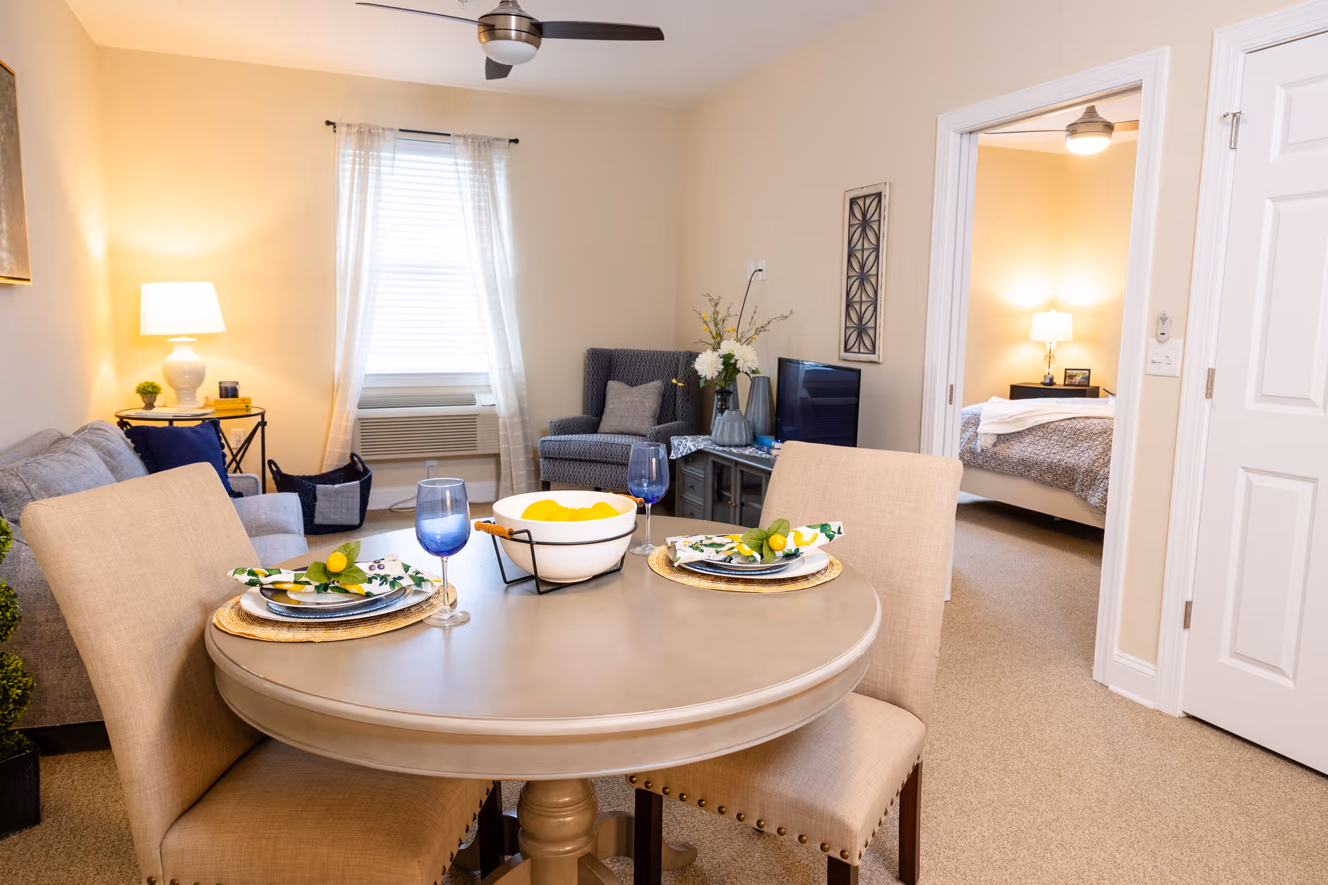 A cozy living and dining area in a senior living facility. The foreground features a round dining table set for two with beige chairs, blue wine glasses, plates, and napkins decorated with lemons. Behind the table is a living room with a gray sofa, a patterned armchair, a TV on a stand, and a window with sheer curtains. A doorway leads to a bedroom with a bed, nightstand, and lamp. The room is warmly lit with neutral walls and carpeted floors.