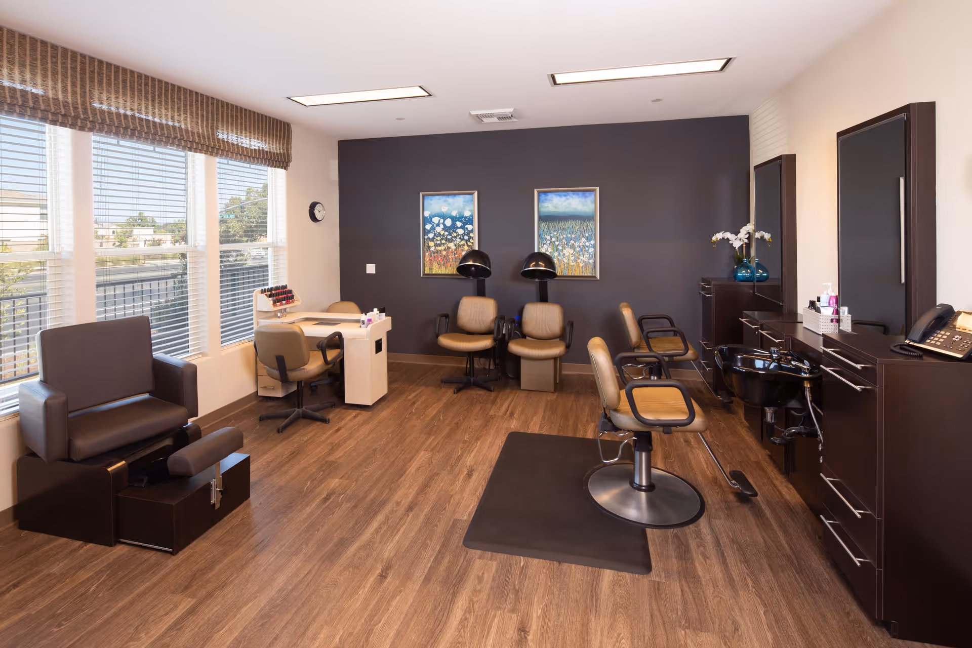 A well-lit salon room with wooden flooring and large windows with blinds. The room contains salon chairs, a pedicure chair, a manicure table, and a hair washing station. The walls are decorated with two framed paintings of floral scenes, and there are modern light fixtures on the ceiling.