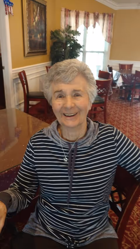A smiling older woman seated at a table in a dining area with chairs, patterned carpet, and a window in the background.