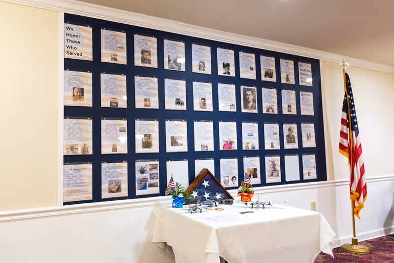 A memorial wall display honoring veterans with multiple framed photos and biographies arranged in a grid on a dark blue background. A table covered with a white cloth is in front of the wall, holding a folded American flag in a triangular case, small American flags, and miniature model airplanes. An American flag on a stand is positioned to the right of the display.