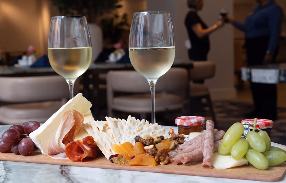 A charcuterie board with grapes, cheese, sliced meats, crackers, dried apricots, nuts, and small jars of jam, accompanied by two glasses of white wine on a table in a dining area with blurred background showing people and chairs.