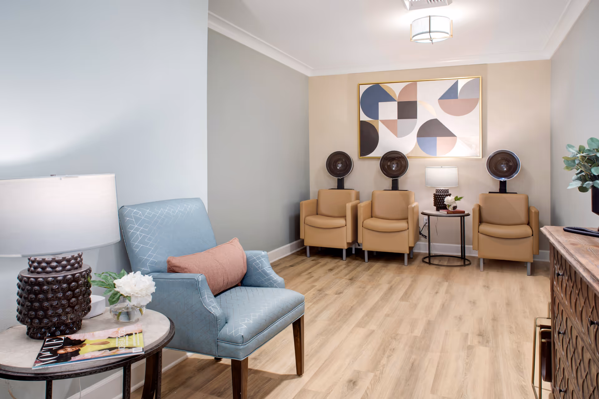 Bright seating lounge with a blue accent chair, three tan armchairs under hair-drying stations, side tables, and abstract wall art.