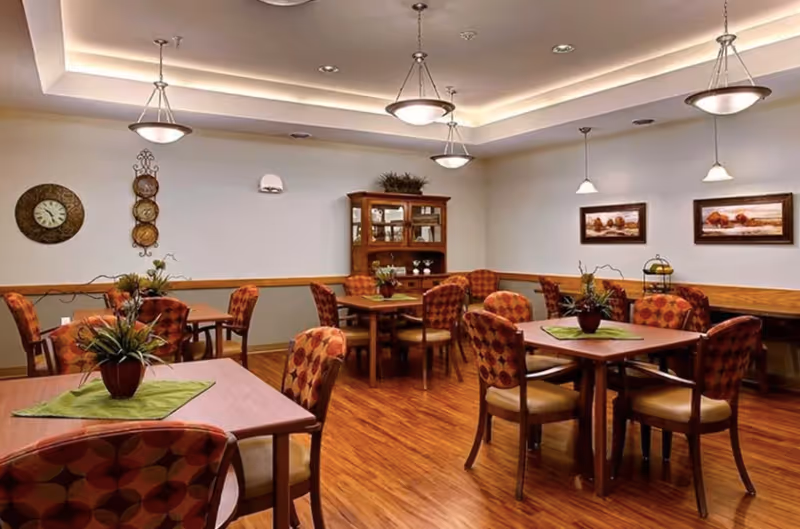 Warmly lit senior living dining room with multiple wooden tables, patterned chairs, potted centerpieces, and framed wall art.