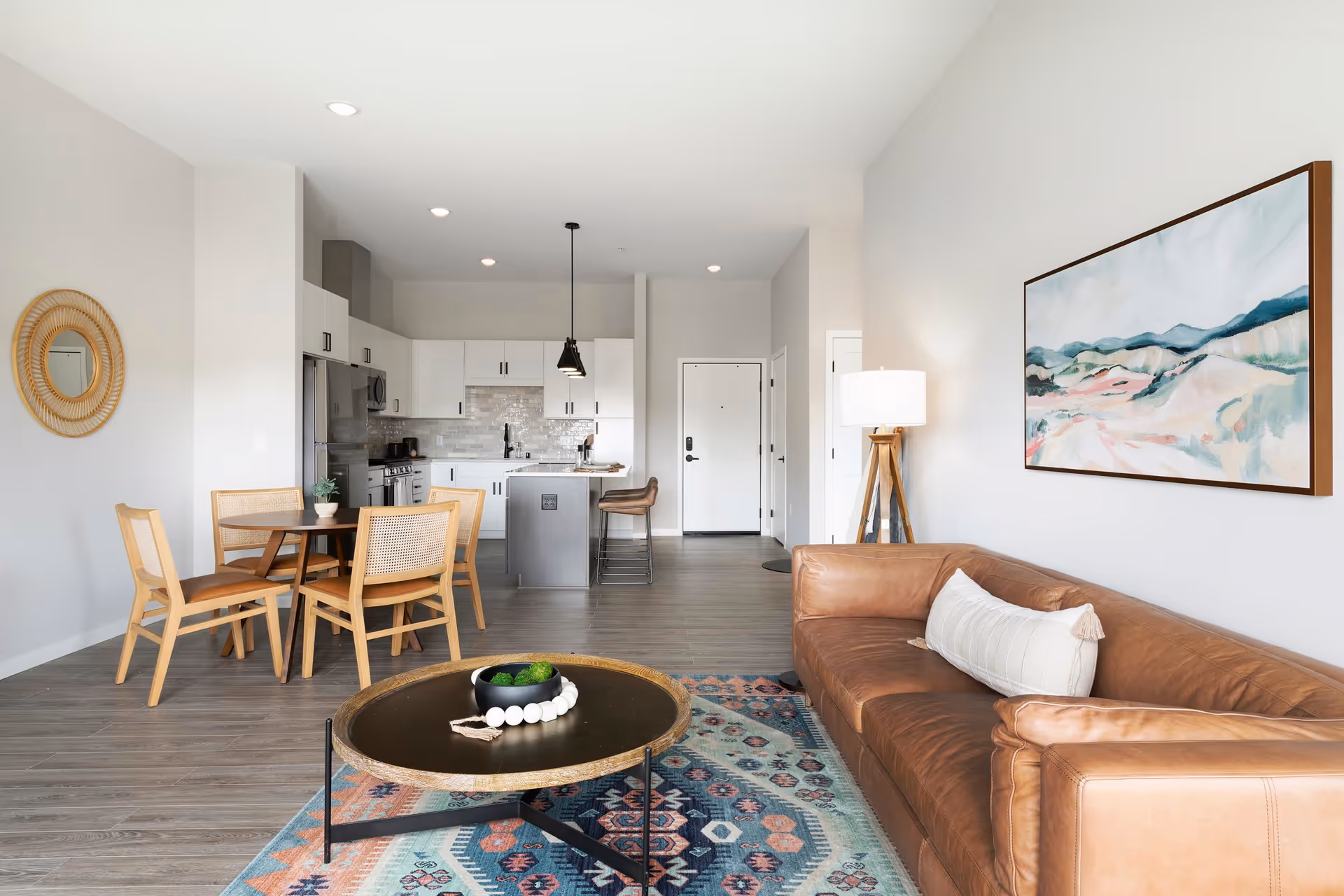 A modern open-concept living space featuring a brown leather sofa with a white pillow, a round coffee table on a colorful patterned rug, a wooden dining table with four chairs, and a kitchen area with white cabinets, stainless steel appliances, and a small island with two bar stools. The walls are light gray with a round mirror and a landscape painting, and there is a tripod floor lamp next to the sofa.
