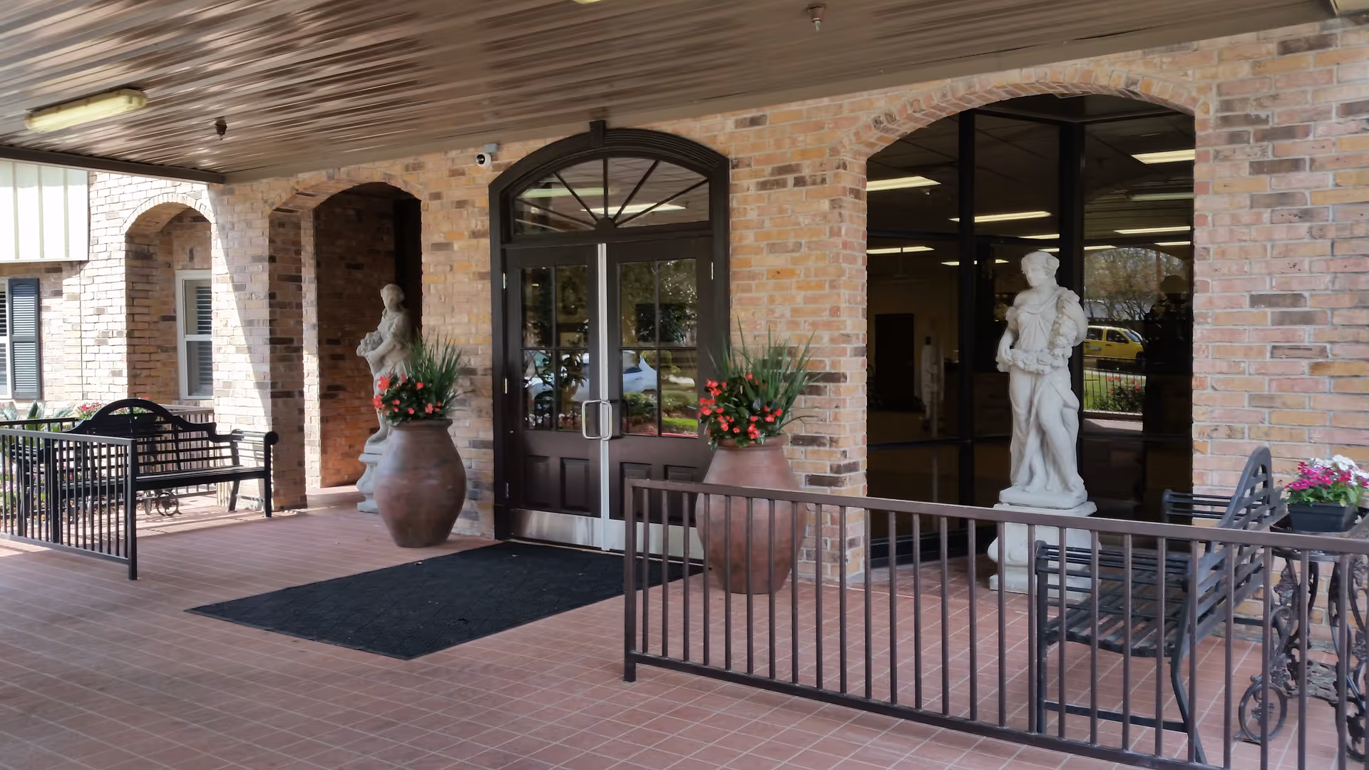 Covered entrance area of a brick building with double glass doors, two large flower pots with red flowers, two white statues, black metal benches, and a black railing.