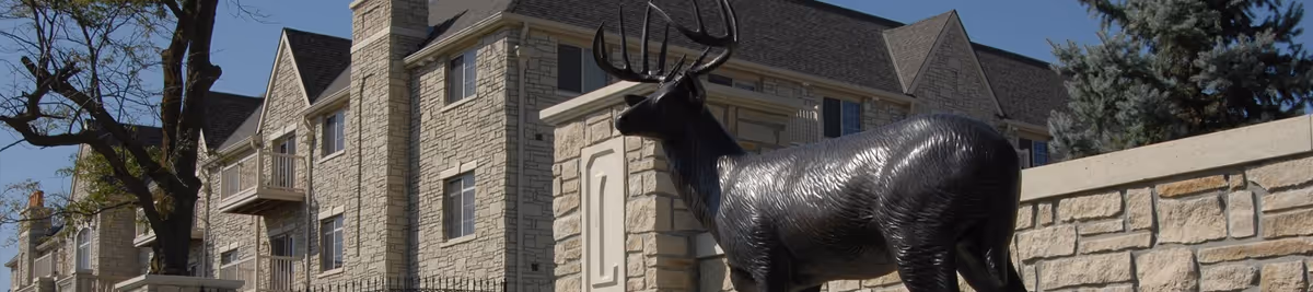 Stone-faced multi-story retirement community building with a life-size elk statue in the foreground and trees.