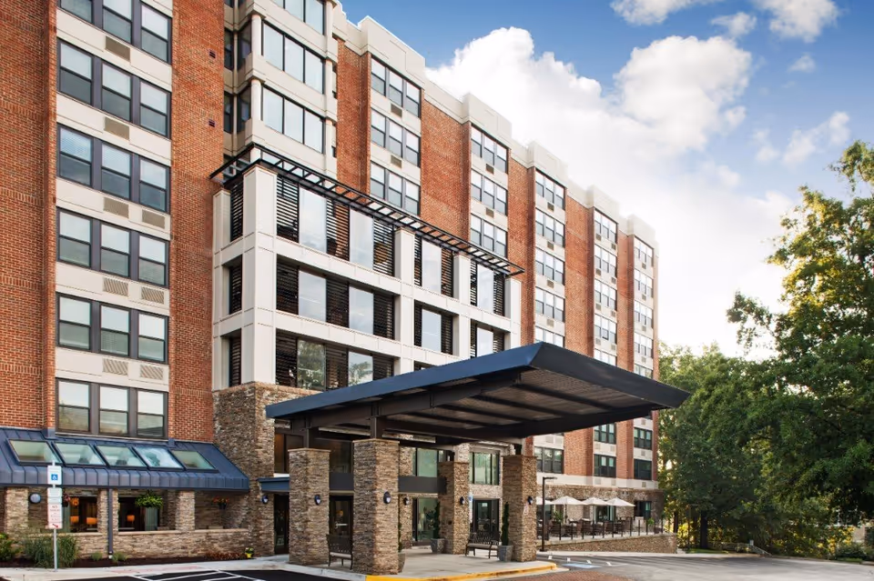 Front entrance of a multi-story brick senior living building with a covered porte-cochère and surrounding trees.