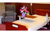 A nursing center bedroom featuring a hospital-style bed with a wooden headboard and footboard, a beige blanket, a bedside table with a flower arrangement in a purple vase, and a comfortable gray armchair in the corner.