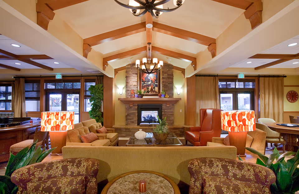 A cozy living room area in Walnut Village Orange County Retirement Community featuring a stone fireplace with a painting above it, surrounded by comfortable seating including sofas, armchairs, and a red leather chair. The room has warm lighting from decorative lamps with patterned shades and a chandelier hanging from a wooden beam ceiling. Large windows with curtains allow natural light to enter, and there are plants adding greenery to the space.