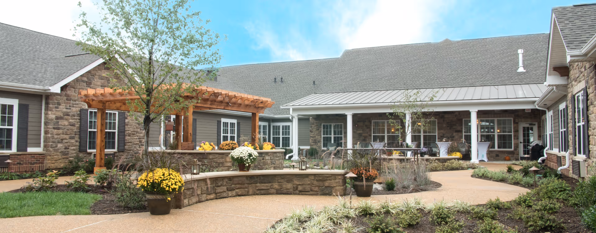 Outdoor courtyard area of a senior living facility with a stone building surrounding it, featuring a wooden pergola, potted yellow and white flowers, small trees, shrubs, and a paved walkway under a blue sky.