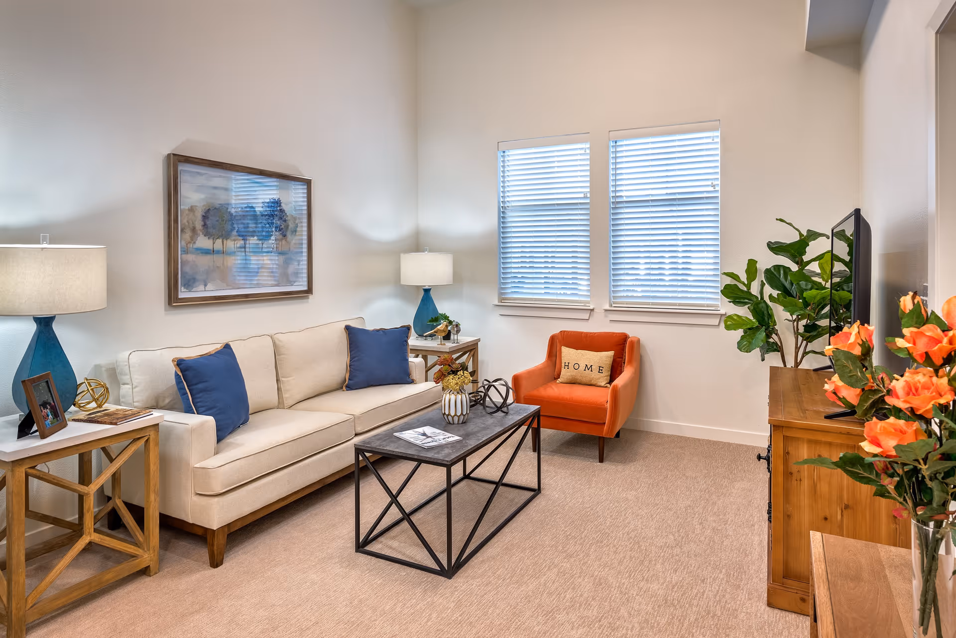 A cozy living room with a beige sofa adorned with two blue pillows, an orange armchair with a 'HOME' pillow, a rectangular coffee table with decorative items, two side tables with blue lamps, a wooden TV stand with a flat-screen TV, and a vase of orange flowers. The room has beige carpet, two windows with white blinds, and a framed painting of trees on the wall.