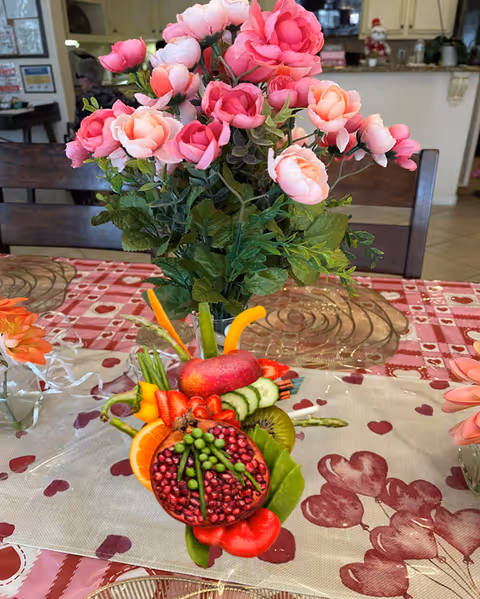A dining table decorated with a bouquet of pink and peach artificial roses in a vase. In front of the flowers, there is an artistic arrangement of various fresh fruits and vegetables including pomegranate, strawberries, kiwi, cucumber, orange slices, and green beans. The table is covered with a tablecloth featuring heart patterns and a transparent decorative runner. The background shows part of a kitchen and dining area with chairs and cabinets.