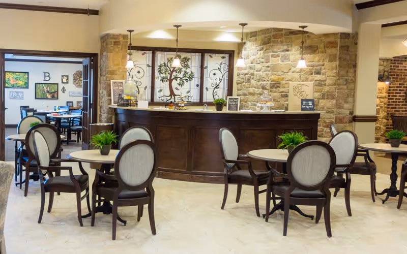 A cozy dining area in a senior living facility featuring round tables with cushioned chairs and small green plants as centerpieces. In the background, there is a curved wooden counter with decorative stained glass windows and stone walls, softly lit by hanging pendant lights. Another dining area is visible through an open doorway to the left.