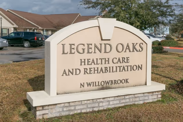 An outdoor stone sign reading 'LEGEND OAKS HEALTH CARE AND REHABILITATION N WILLOWBROOK' in front of a healthcare building and parking lot.