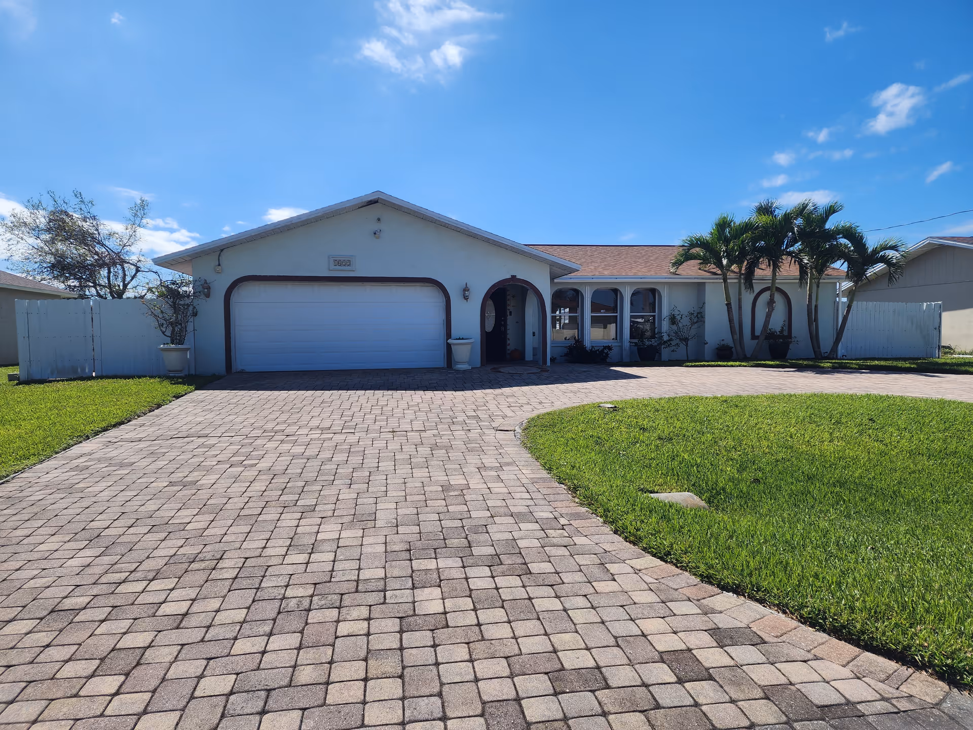 Front exterior view of a single-story house with a large paved driveway, a two-car garage, and a well-maintained lawn with palm trees and other plants under a clear blue sky.