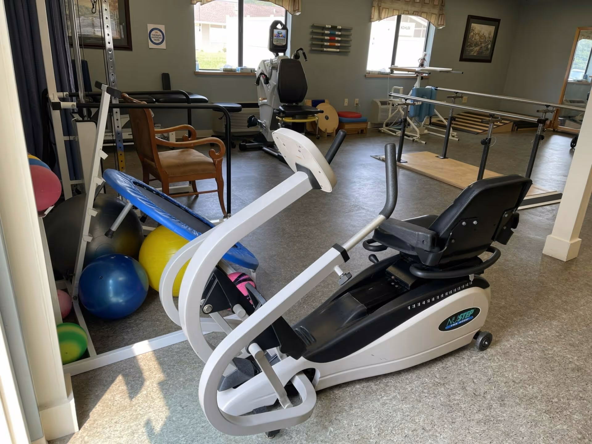 A physical therapy or exercise room with various equipment including a recumbent exercise bike, parallel bars for walking support, exercise balls, a small trampoline, and a wooden chair. The room has windows letting in natural light and framed pictures on the walls.