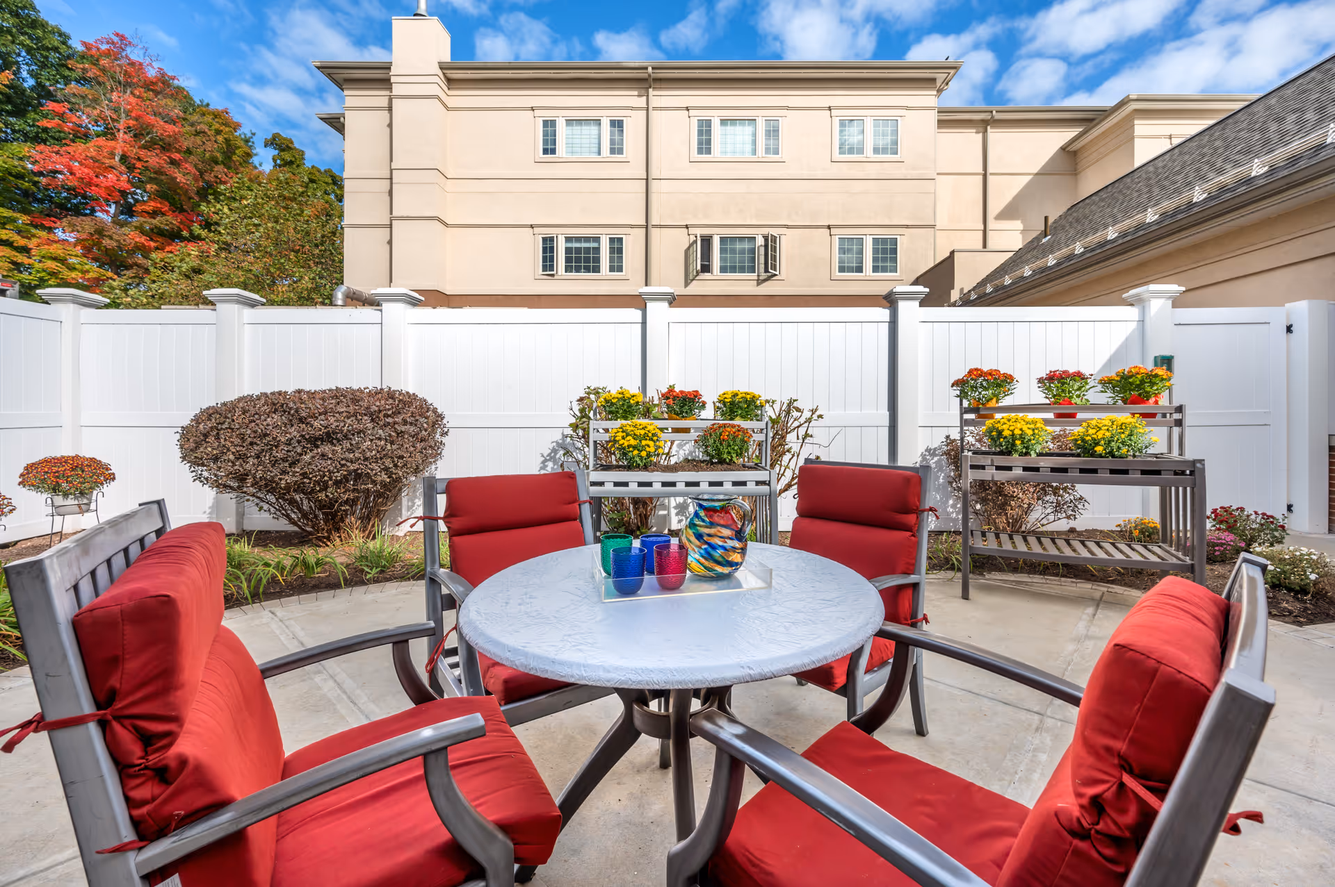 Outdoor patio area with a round table covered with a white tablecloth and surrounded by four chairs with red cushions. On the table, there is a colorful glass pitcher and four colorful glasses. Behind the table, there are two metal plant stands with yellow, orange, and red flowers. The area is enclosed by a white fence, and a beige building with multiple windows is visible in the background under a partly cloudy blue sky.