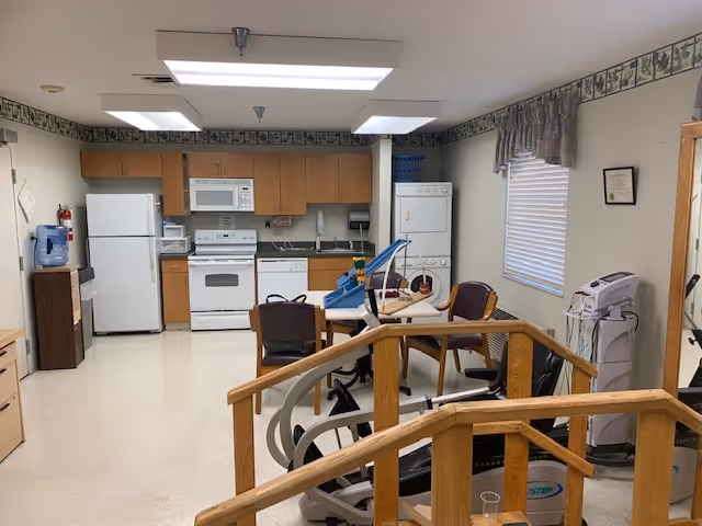 Interior view of a multipurpose room in a senior living facility featuring a kitchen area with white refrigerator, stove, microwave, and dishwasher. In the foreground, there is exercise equipment and a table with chairs. The room has fluorescent lighting, a window with blinds and a valance, and light-colored walls with a decorative border near the ceiling.
