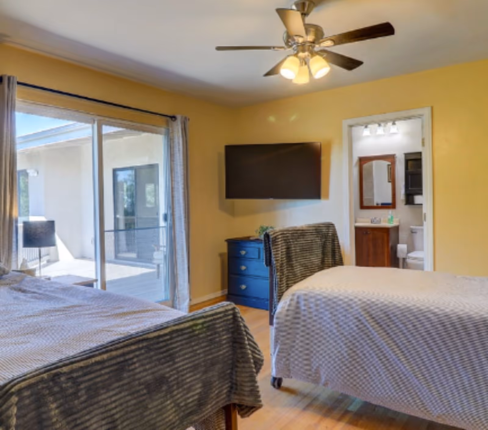A bedroom with two beds covered in gray and white bedding, a ceiling fan with lights, a wall-mounted flat-screen TV, a blue dresser, and a sliding glass door leading to an outdoor patio. A bathroom with a sink, mirror, and toilet is visible through an open door.