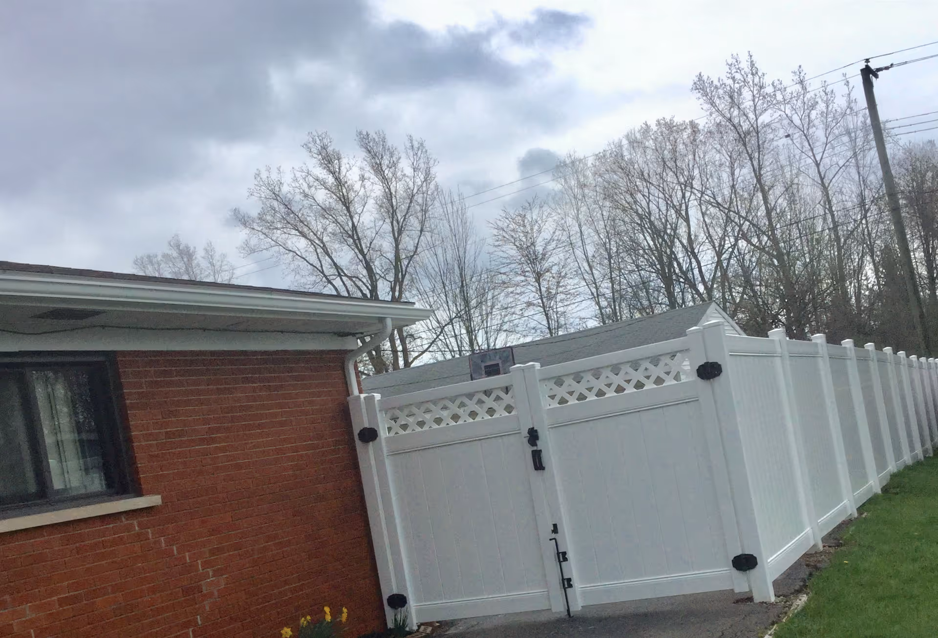 Outdoor view of a brick building with a white vinyl fence and gate. There are leafless trees in the background under a cloudy sky.