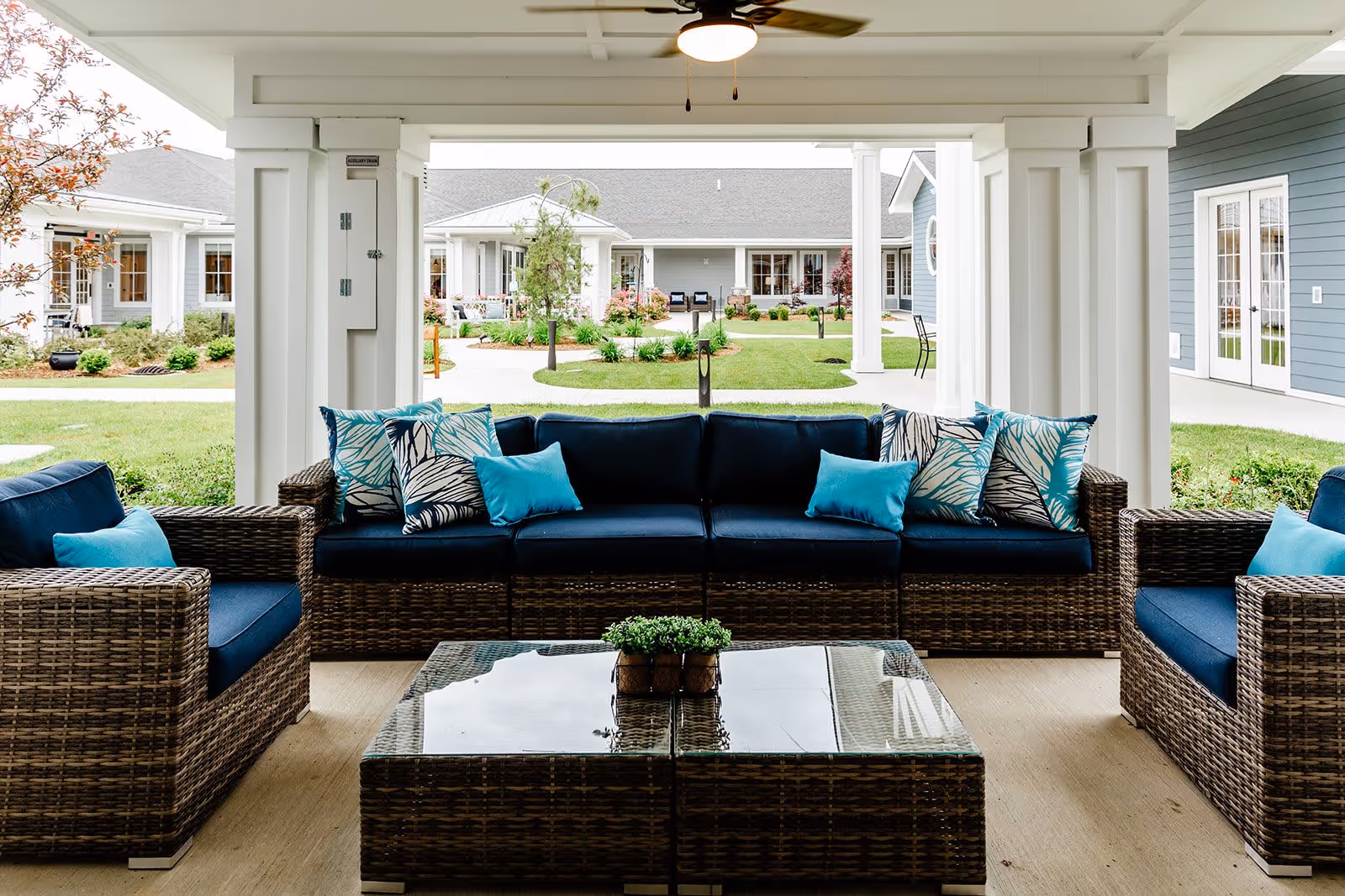 Outdoor covered seating area with wicker furniture including a sofa and two armchairs with blue cushions and decorative pillows. A glass-top wicker coffee table with a small green plant is in the center. The area overlooks a landscaped courtyard with green grass, shrubs, and pathways, surrounded by light blue buildings with white trim.