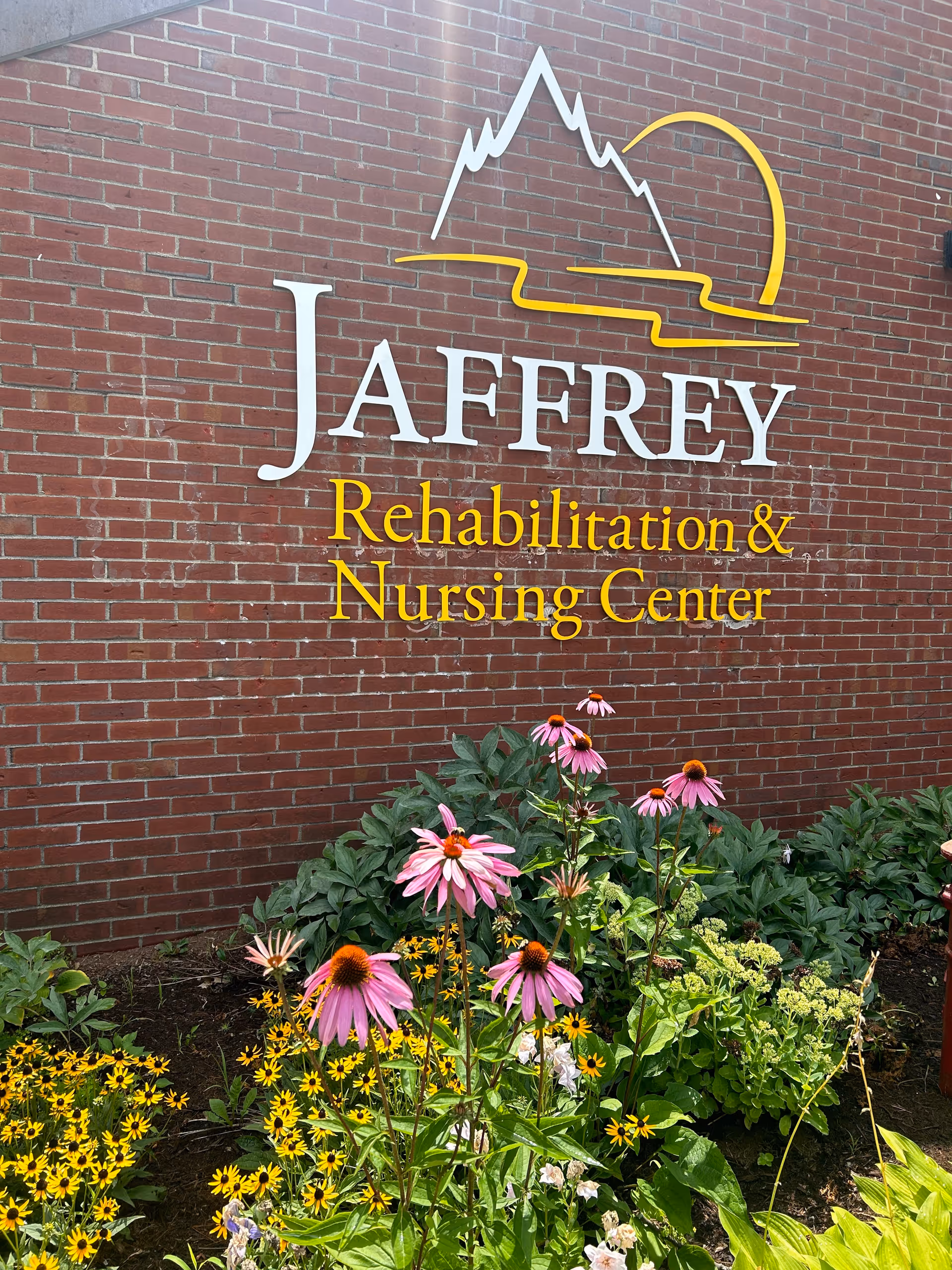 Sign reading 'Jaffrey Rehabilitation & Nursing Center' on a red brick wall above a flower bed with pink coneflowers.