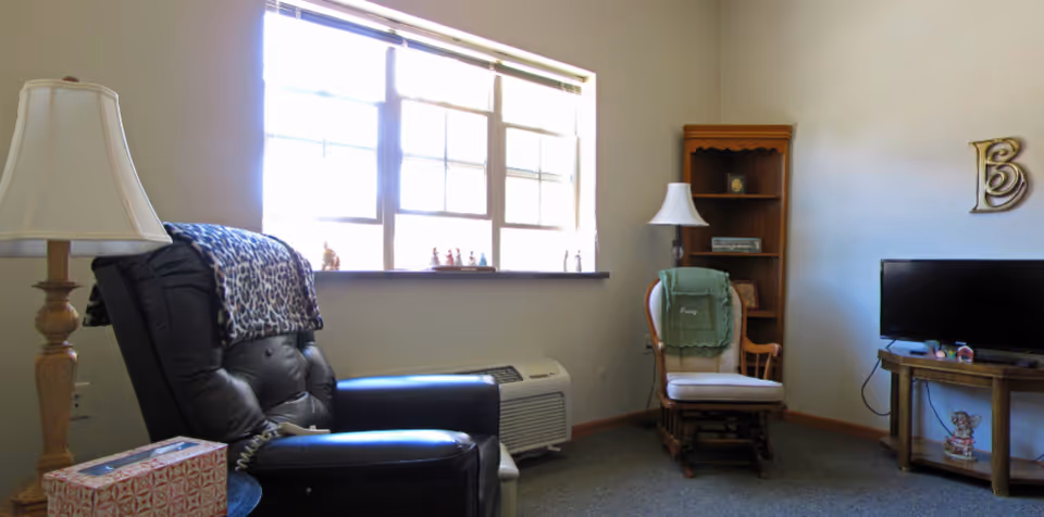 A cozy living room area with a large window letting in natural light. The room features a black leather recliner with a leopard print blanket draped over it, a wooden rocking chair with a green throw blanket, a wooden bookshelf with a lamp on top, and a small wooden TV stand holding a flat-screen television. The walls are light-colored, and there is a decorative letter 'B' mounted on the wall.