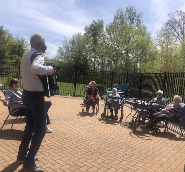 An outdoor patio area with a group of elderly people seated in chairs and wheelchairs arranged in a semicircle. A man wearing a face mask is standing and playing an accordion for the group. Trees and a black metal fence are visible in the background under a partly cloudy sky.