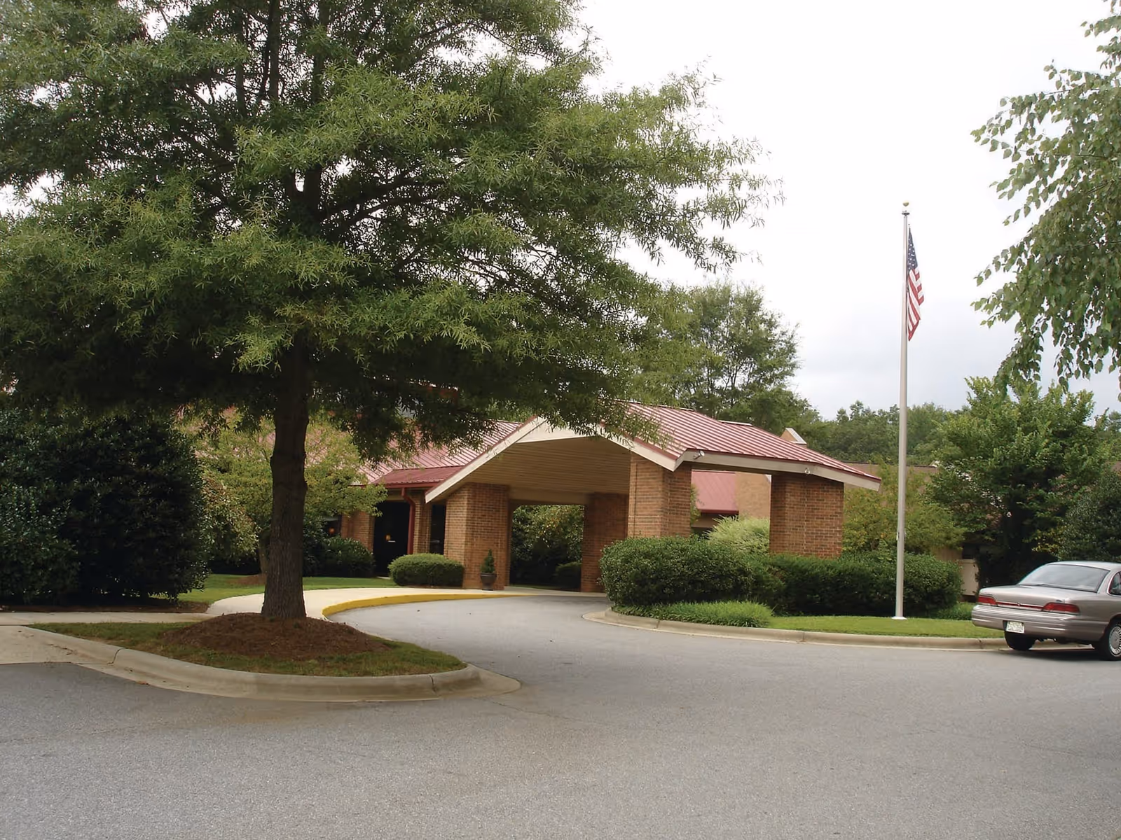Entrance area of a facility with a covered driveway supported by brick pillars, surrounded by green trees and bushes, an American flag on a flagpole, and a parked car on the right side.