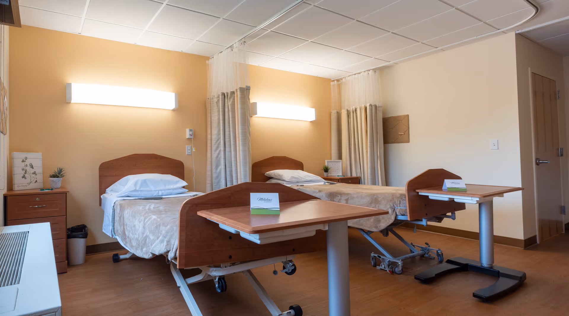 A nursing facility room with two hospital-style beds separated by privacy curtains. Each bed has a beige blanket and white pillows. There are two overbed tables, one in front of each bed, and a small wooden nightstand with a plant and decorative item. The walls are painted light beige, and there are bright wall-mounted lights above each bed.