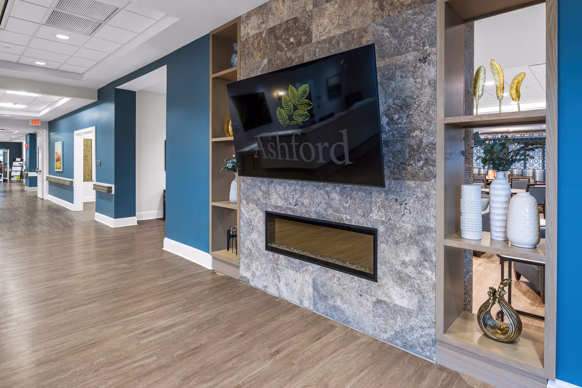 Interior hallway of The Ashford of Beavercreek facility featuring wood flooring, blue and white walls, a mounted flat-screen TV displaying the Ashford logo above a modern electric fireplace, and decorative shelving with vases and ornamental items. The hallway extends with handrails and framed artwork on the walls.