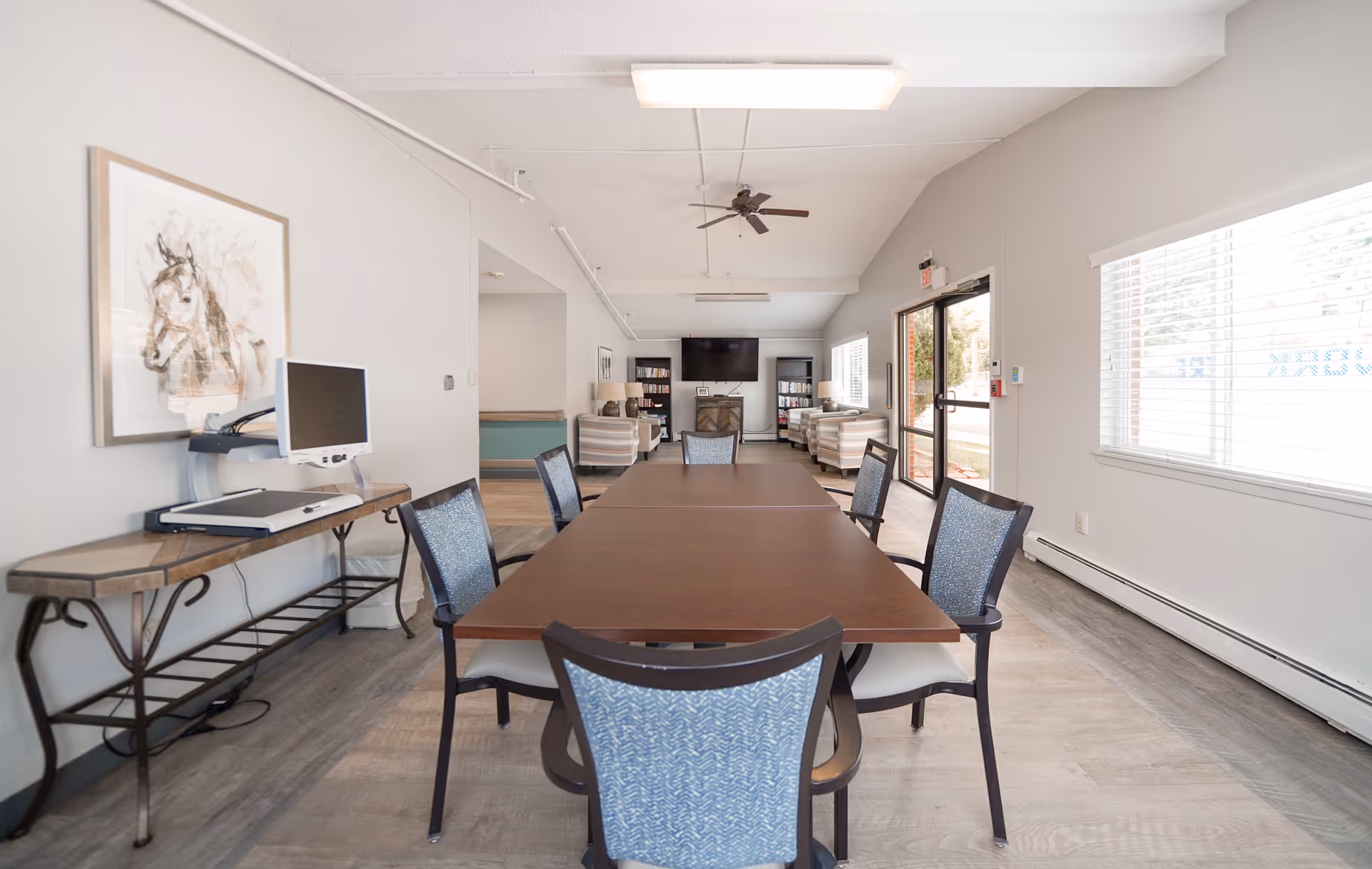 A bright and spacious common area with a long wooden table surrounded by six chairs with blue patterned upholstery. To the left, there is a table with a computer monitor and keyboard. The room has light-colored walls, large windows with blinds on the right, and a ceiling fan. In the background, there are comfortable armchairs, bookshelves, and a wall-mounted TV above a fireplace. The entrance door is visible on the right side.