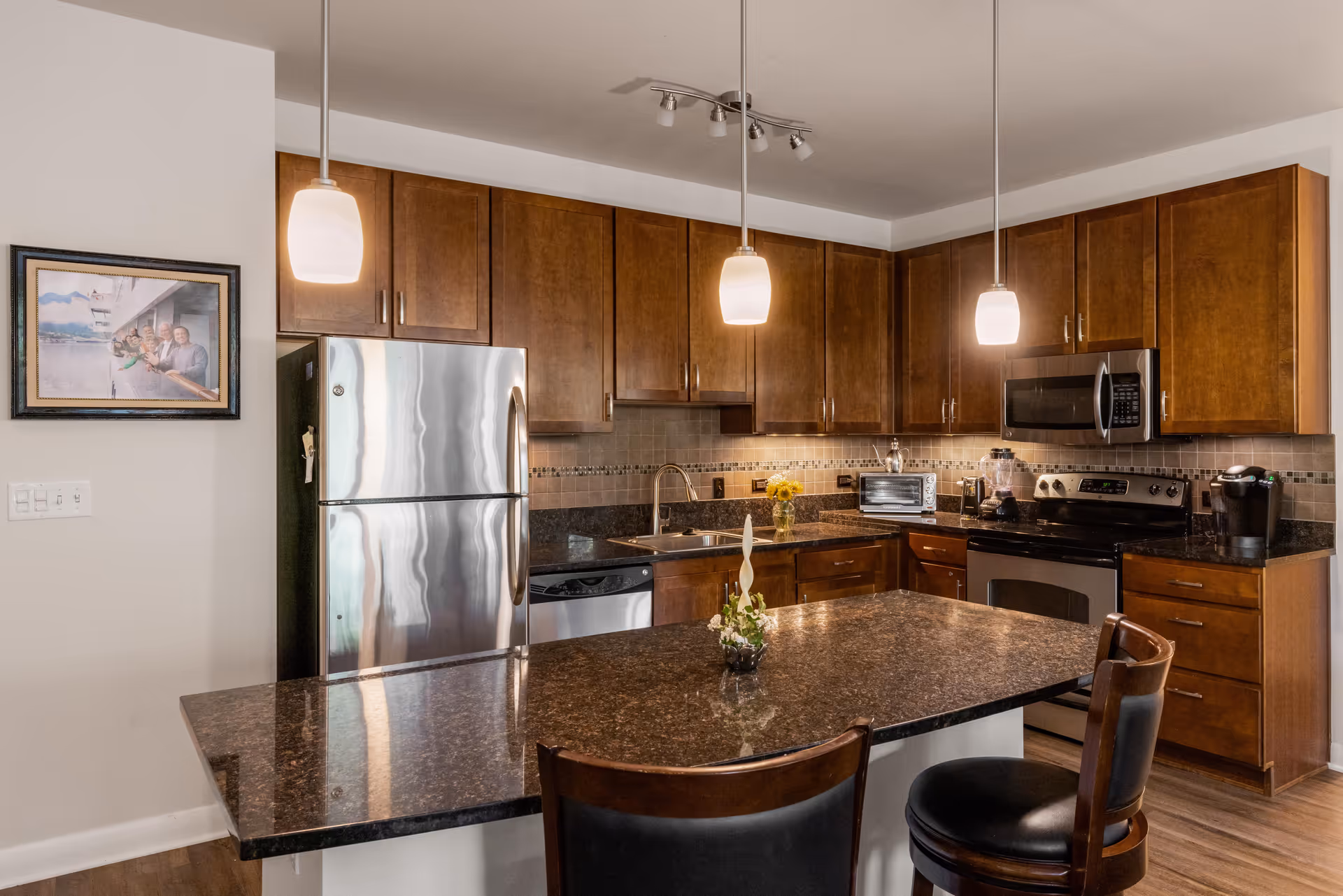 Modern kitchen with dark granite countertops, wooden cabinets, stainless steel refrigerator, dishwasher, microwave, and stove. Two pendant lights hang over a kitchen island with two chairs. A small vase with flowers is on the island, and a framed photo is on the wall to the left.