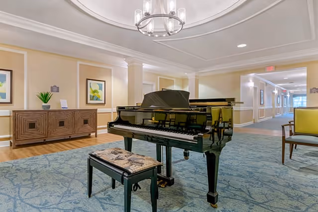 A spacious and well-lit common area featuring a black grand piano with a cushioned bench on a blue patterned carpet. The room has beige walls with white trim, two framed artworks, a wooden sideboard with a small plant and a card, and a modern chandelier hanging from the ceiling. A hallway extends in the background with additional seating and lighting.