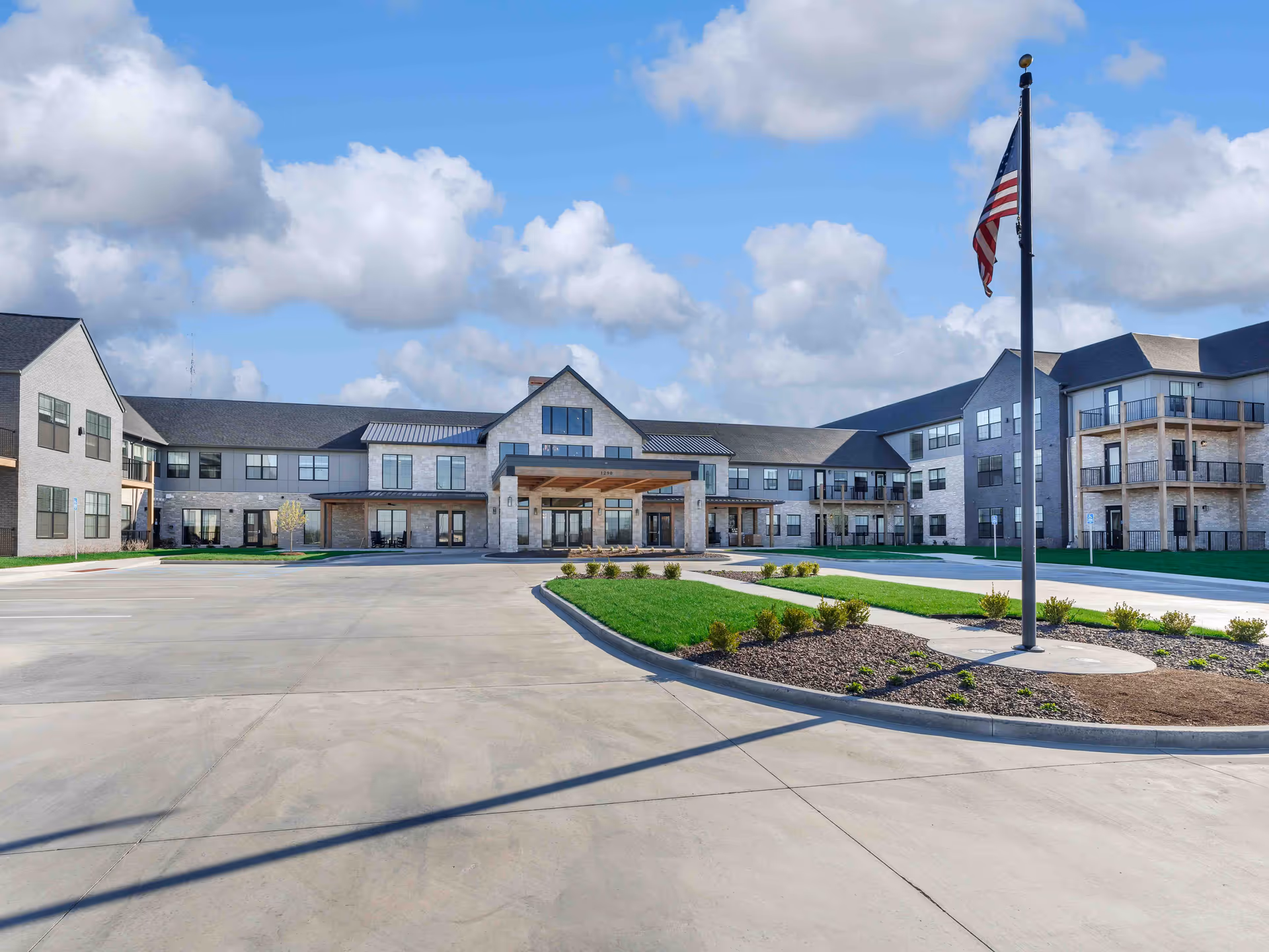Front exterior view of Cedarhurst Senior Living of Wentzville building with a large driveway, landscaped areas, and an American flag on a flagpole under a partly cloudy sky.
