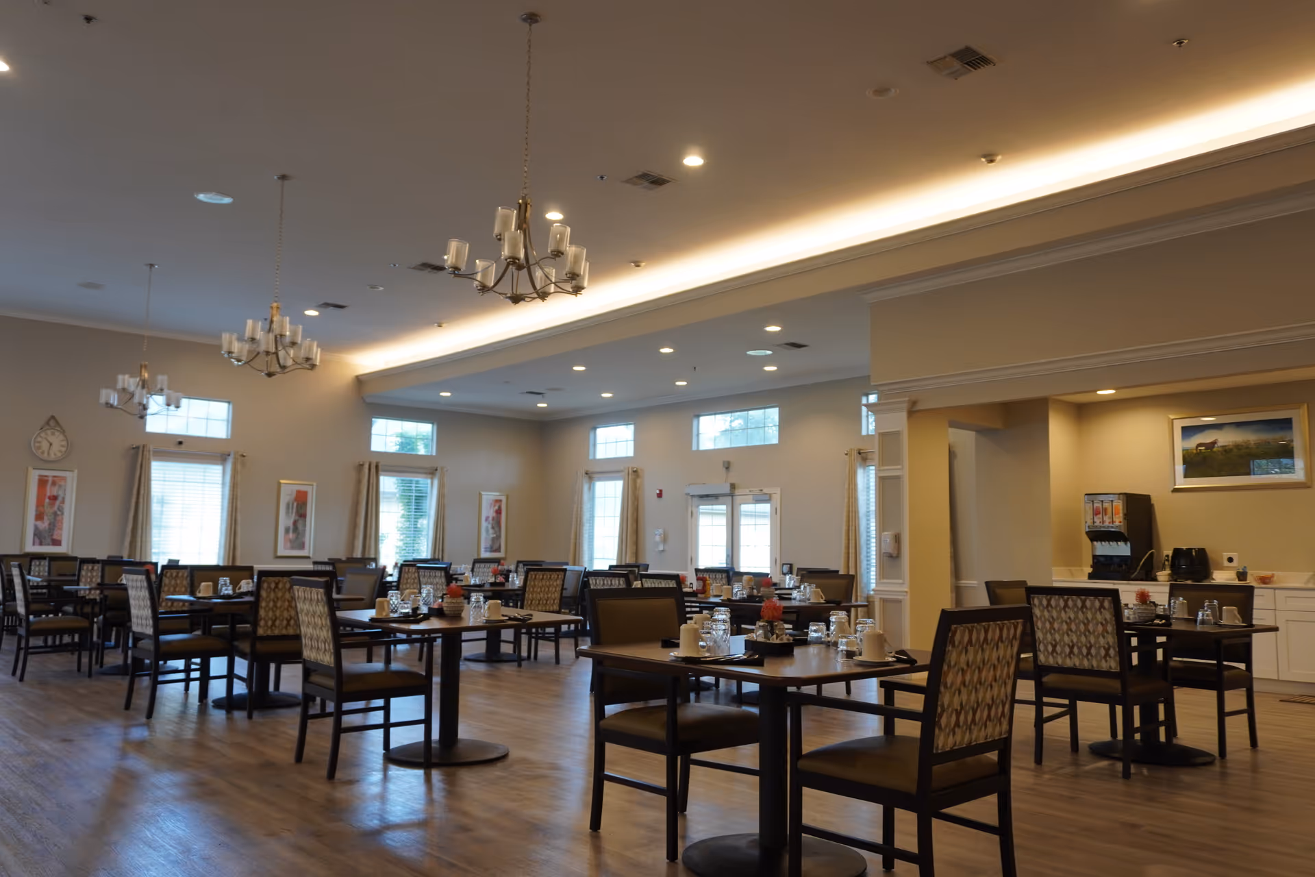 A spacious dining room with multiple tables and chairs arranged neatly. Each table is set with cups, glasses, and small decorative centerpieces. The room features large windows with curtains, chandeliers hanging from the ceiling, and a beverage station with a drink dispenser and air fryer in the corner.
