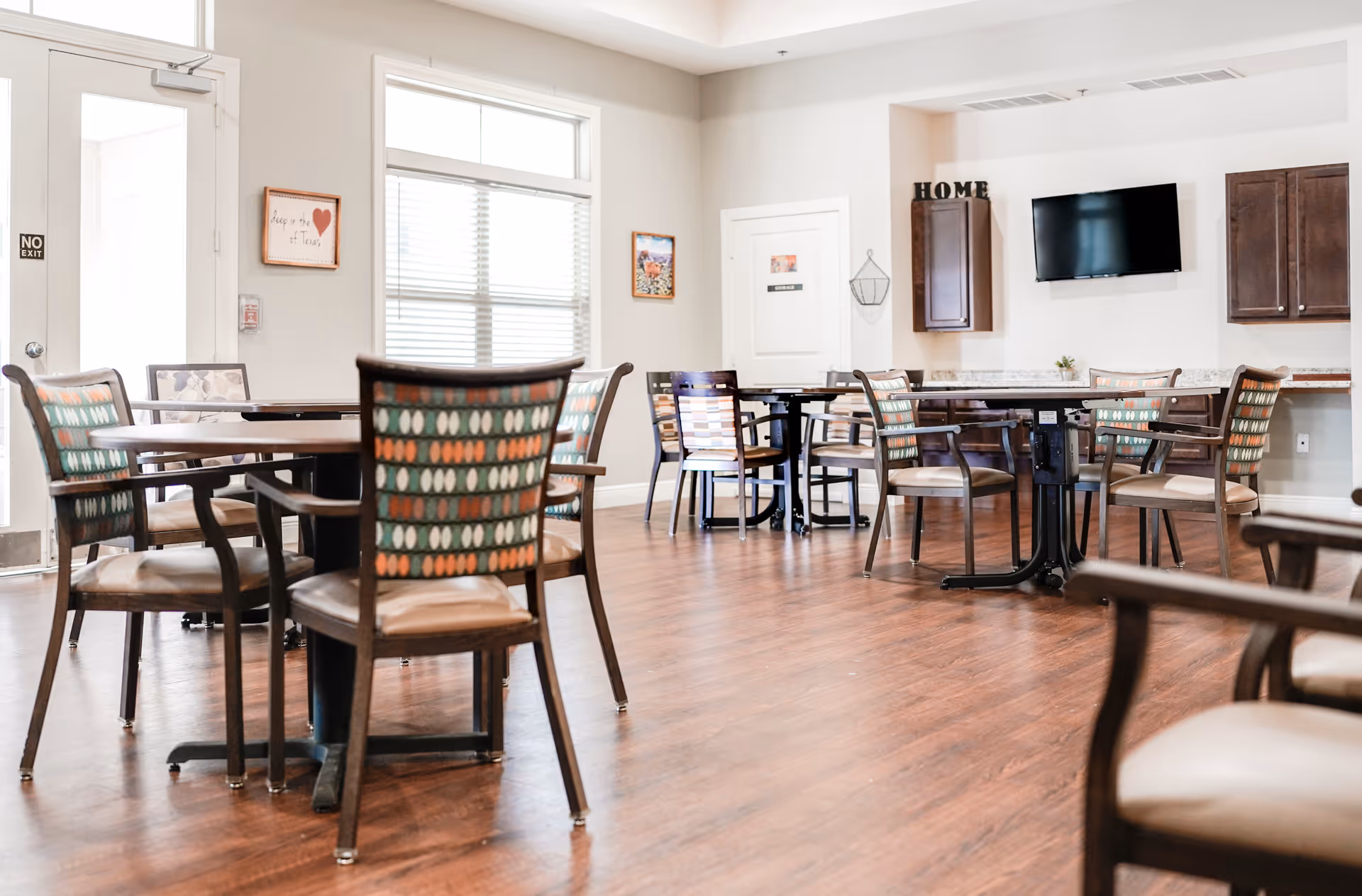 Bright communal dining area with round tables, patterned chairs, and a wall-mounted TV.