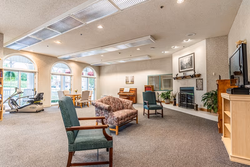 A spacious senior living facility common area with carpeted floor, featuring a floral-patterned sofa, green armchairs, a piano against the far wall, a fireplace with framed artwork above it, and a television on a wooden stand. Large arched windows and doors allow natural light to enter, and exercise equipment is visible near the windows.
